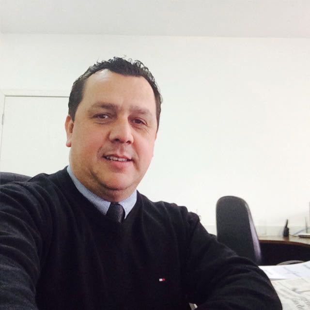 Foto em ambiente de escritório com um homem de terno escuro, sorrindo e sentado, em frente a uma mesa com documentos.