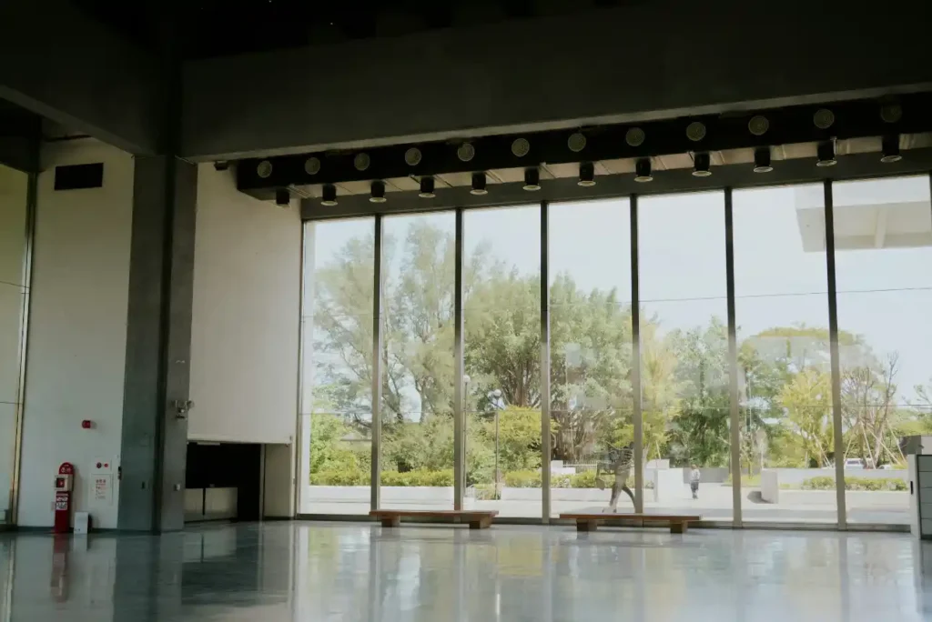 Vista interna de um salão com piso de concreto e grandes portas de vidro do chão ao teto, com jardim e árvores ao fundo sob luz natural.