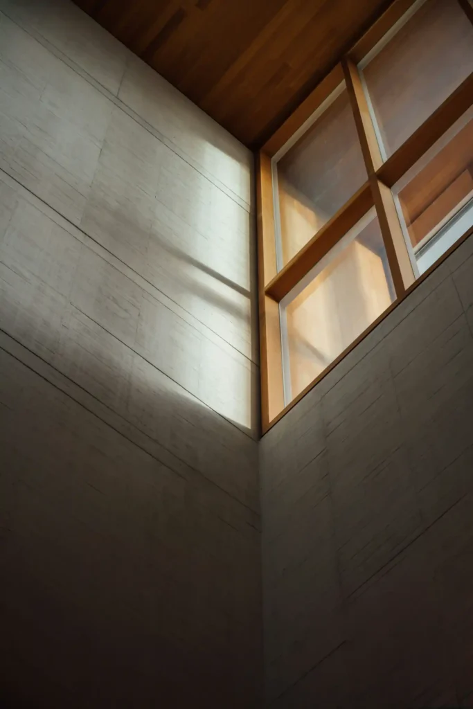 Detalhe de arquitetura interior com paredes de madeira e revestimento em tom cinza, além de treliças e áreas de luz natural formando um ângulo no teto. Linhas e sombras geométricas.