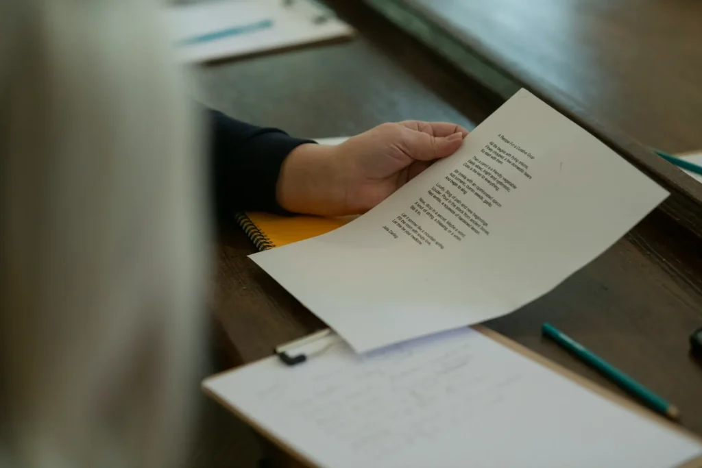 Pessoa em escritório com uma mão segurando uma folha de papel com texto impresso, sobre uma mesa de madeira, ao lado de cadernos e lápis, em ambiente de trabalho.