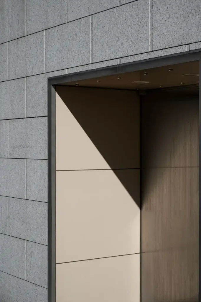 Entrada de edifício com fachada de concreto cinza e moldura metálica, mostrando uma área interna com painel de vidro e sombreamento em diagonal.