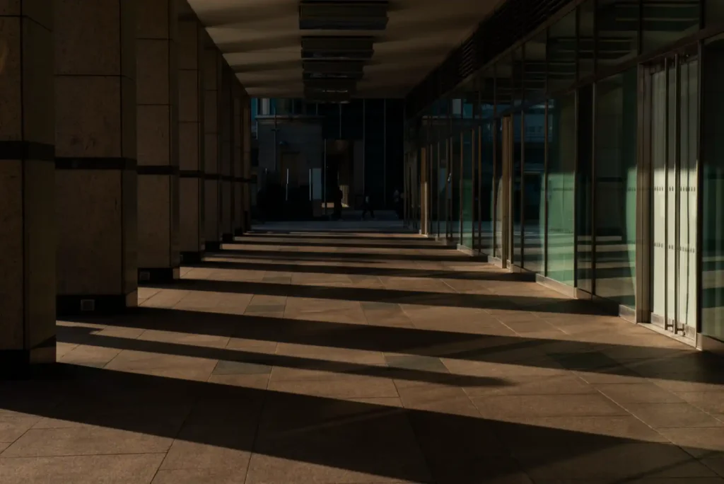 Corredor moderno de prédio com paredes de pedra, piso de placas e grandes portas de vidro refletindo a luz do fim da tarde, criando sombras longas.