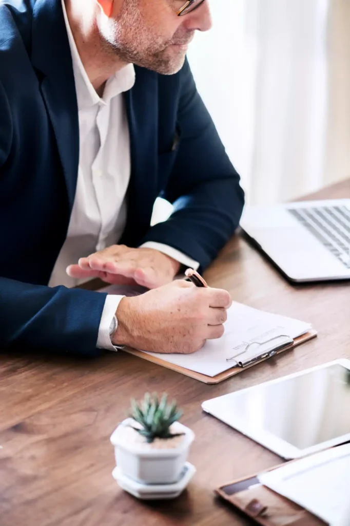 Homem de terno em reunião empresarial sentado à mesa, analisando documentos e anotando informações com caneta, ao lado de notebook e pastas.
