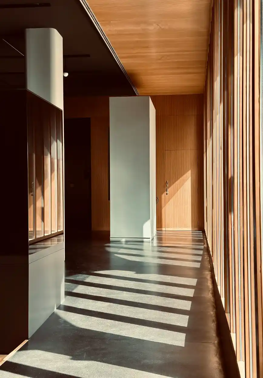 Corredor moderno com paredes revestidas em madeira e painéis claros, iluminação natural do lado e faixas de luz projetadas no piso, em arquitetura contemporânea.