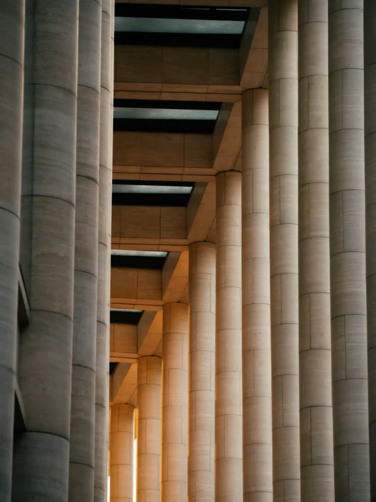 Vista em perspectiva de colunas e arcos monumentais de pedra, com luz quente atravessando o espaço entre as estruturas e criando um efeito de profundidade.