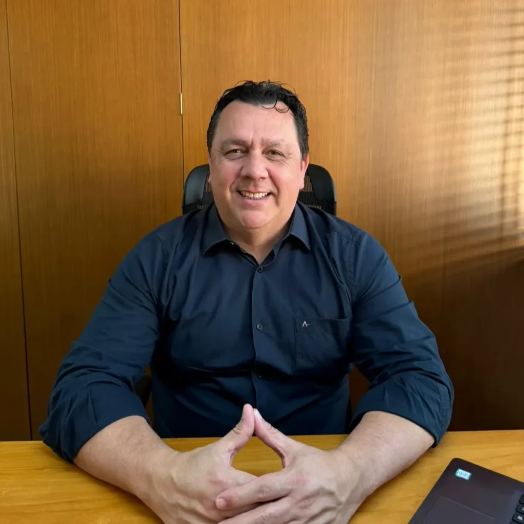 Adinan, da A Chave Leilões, sorrindo sentado à mesa de escritório, com camisa azul-marinho e as mãos unidas sobre a mesa de madeira.